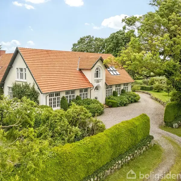 En stor, hvid villa med rødt tegltag står omgivet af frodig vegetation og grusvej. Huset er omgivet af hække, træer og en velplejet græsplæne i naturskønt område.