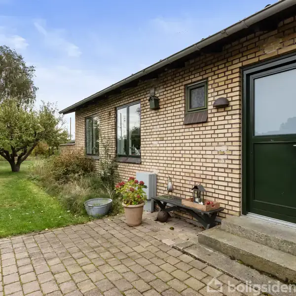 Hus med murstensfacade og mørkegrøn dør, flankeret af blomsterkrukker og bænk. Foran er en flisebelagt terrasse, og haven har græsplæne og træer i baggrunden under en blå himmel.