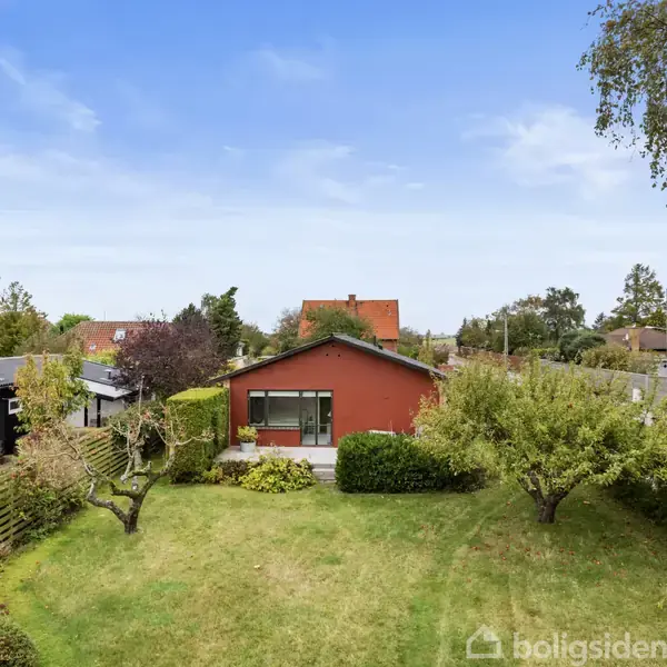 Et rødt hus står stille i en velplejet have. Omkring huset er der grønne buske, træer og græs, med en klar blå himmel over det hele.