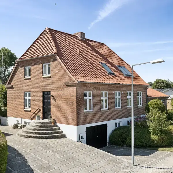 Et rødt murstenshus med rødt tegltag står på en hjørnetomt. Rund trappeopgang leder til hoveddør. Omgivet af hæk og belægningssten. Lampestolpe ses ved fortovet.