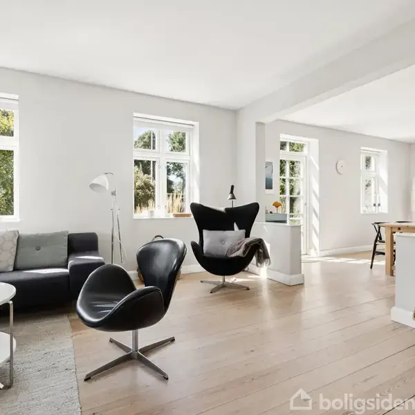 En moderne stue indeholder sorte stole og sofa, arrangeret omkring et lille rundt bord. Rummet er lyst med store vinduer og åbner op til et køkkenområde.