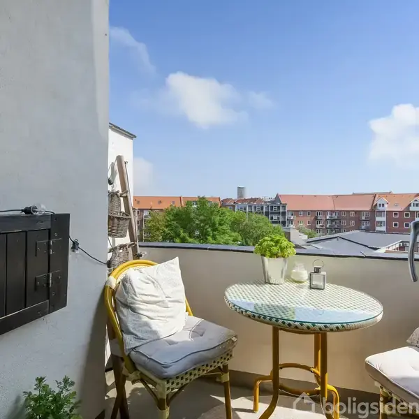 Et lille balkonbord med to stole står i et hjørne med udsigt over en by. Grønne planter står på bordet, og lys fylder scenen under en klar blå himmel.