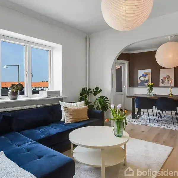 En stue med en mørkeblå sofa ved siden af et rundt hvidt sofabord med blomster i en vase. I baggrunden ses en spisestue med seks stole og dekorative billeder på væggen.
