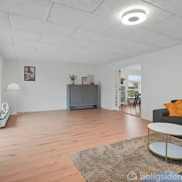 Stue med sofa og sofabord på gulvtæppe. En reol står ved væggen nær en døråbning. Store vinduer lukker lys ind i det rummelige, minimalistisk indrettede rum.