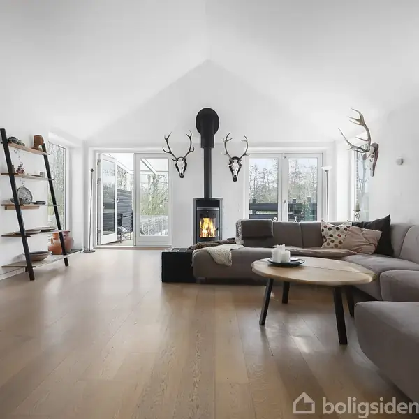 En moderne stue med en grå hjørnesofa står foran en brændeovn, hvor flammerne brænder livligt. Naturtro dekoration med hjortegevirer pryder væggene. Rummelige vinduer giver lys og udsigt.