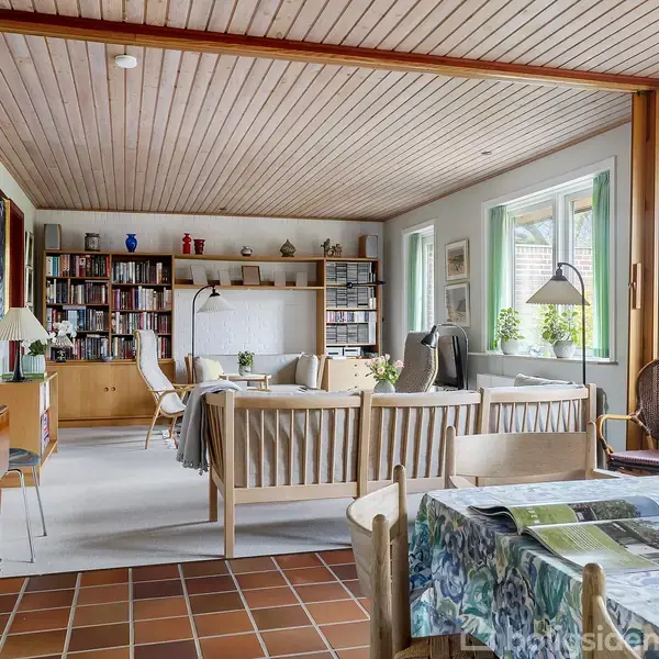 Stueområde med trægulv og træloft, indrettet med lænestole og sofabord. Et bord med dug står nær vinduer. Baggrunden viser bogreoler og et skrivebord. Grønne planter og lamper udstråler hygge.