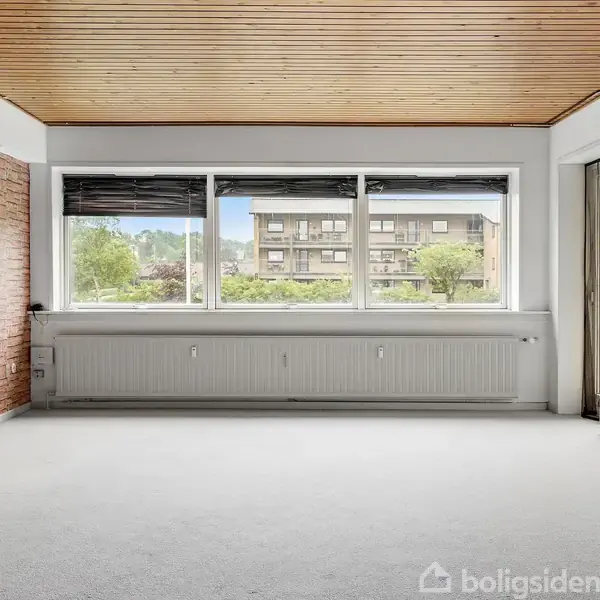 Et tomt rum med tæppebelagt gulv har en væg med murstensdesign og store vinduer. Udenfor kan man se et lejlighedskompleks og træer. Loftet er af træ.