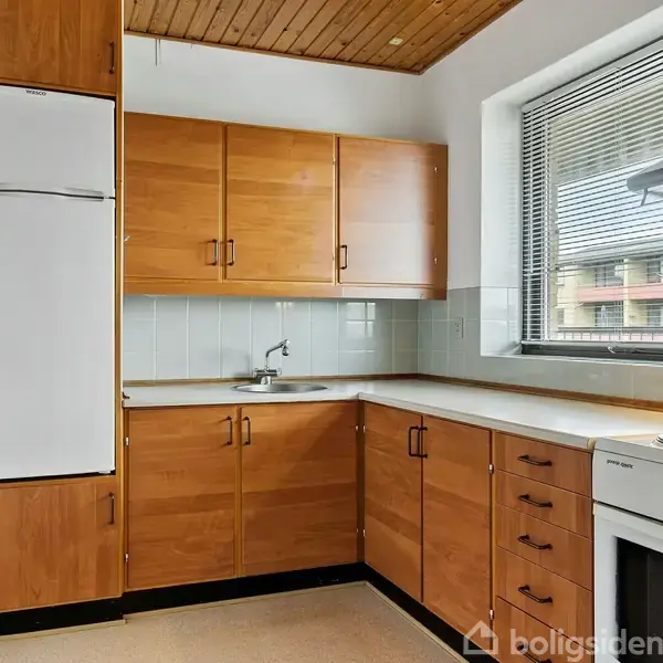 Køkken med træskabe og hvidevarer. Et vindue med persienner til højre giver udsigt til en boligbygning. Det er velorganiseret med et komfur og vask under et træloft.