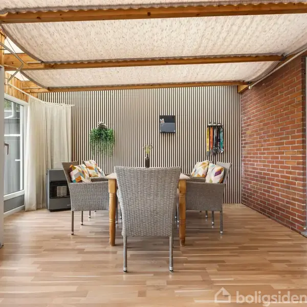 Stole og bord står i et lyst, overdækket terrasseområde med trægulv. Omkring dem er glasdøre og mursten. En plante og pynt hænger på væggen.