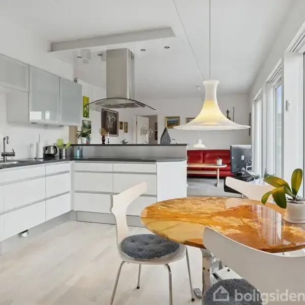 Et moderne køkken-alrum har et rundt bord med en gylden bordplade, omgivet af stole, med en stor lampe hængende over. Store vinduer giver udsigt til en terrasse.