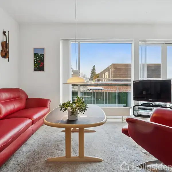 Rød sofa og lænestol står omkring et træbord med en plante, under en hængende lampe, i en lys stue med store vinduer og udsigt til bygninger udenfor.