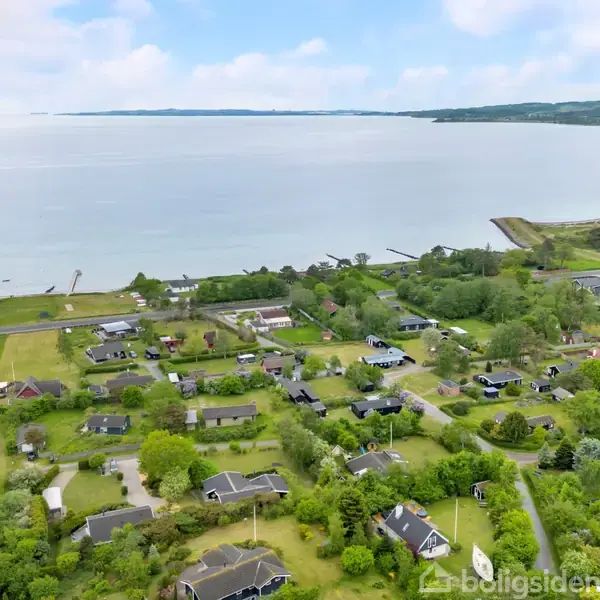 En kystnær landsby, hvor huse spredt blandt grønne træer ses fra luften. Rolige bølger strækker sig mod horisonten under en delvist skyfri himmel.