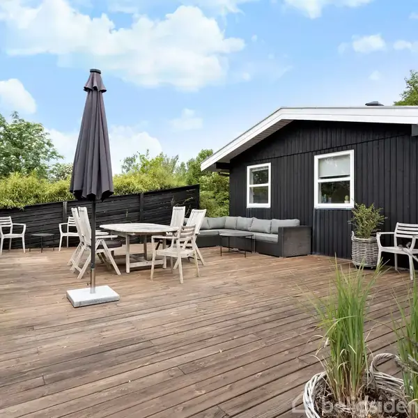 Terrassen har et trædæk med et mørkt hegn, hvor der står et bord med stole og en parasol. En sort træbygning med vinduer står i baggrunden, omgivet af grønne planter.