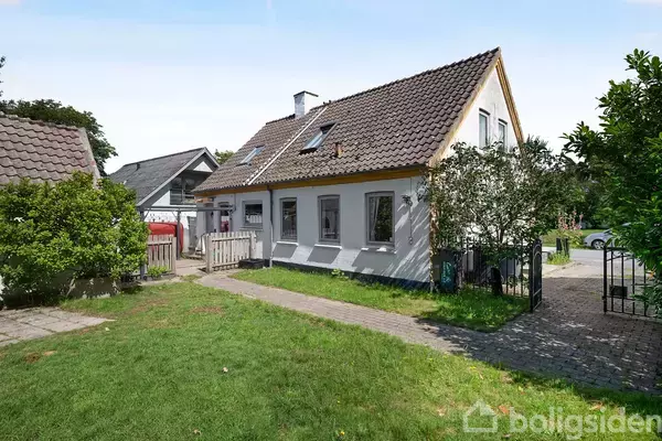 Et hvidt murstenshus med brunligt tegltag står i en grøn have med stier og buske. Ved siden af huset er der en indkørsel og en lille terrasse.