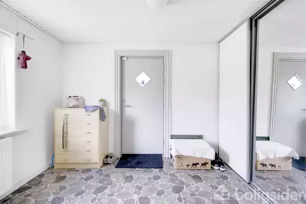 En entré med en kommode til venstre, en lukket dør foran, og spejle til højre. Gulvet består af stenfliser, og en papkasse står ved skoskabet.