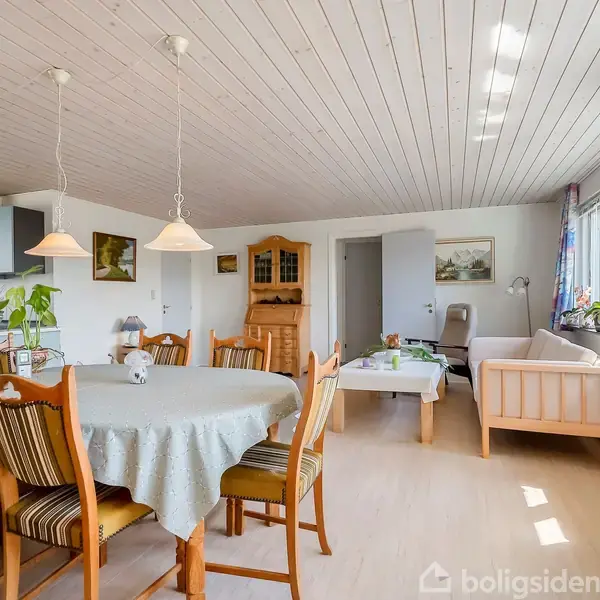 Et spisebord med seks stole er centralt placeret i en lys stue. Sofaen står ved vinduet, og lysekroner hænger over bordet. Malerier pryder væggene.