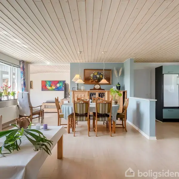 Stue med spisebord, omgivet af stole, står i centrum, belyst af lamper. Konteksten er en lys lejlighed, med planter og køkkenområde i baggrunden.