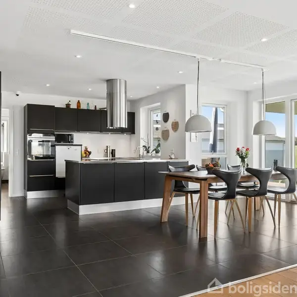 Sort køkken med høje skabe og en kogeø, opdækning på spisebord med blomsterdekorationer i en moderne, lys spisestue, store vinduer med udsigt til haveområde.