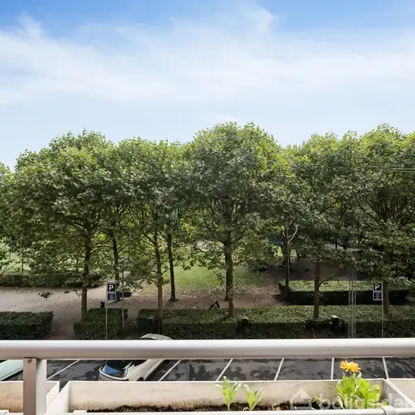 Balkon med udsigt over en parkeringsplads, hvor biler holder parkeret. Træer danner en grøn baggrund under en blå, let overskyet himmel.