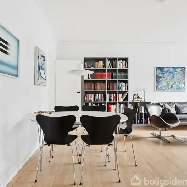 Et moderne spisestueområde med sort-hvide stole og et rundt bord. Bagved ses en bogreol med bøger. En sort lædersofa og moderne kunst hænger på væggene i et lyst rum.