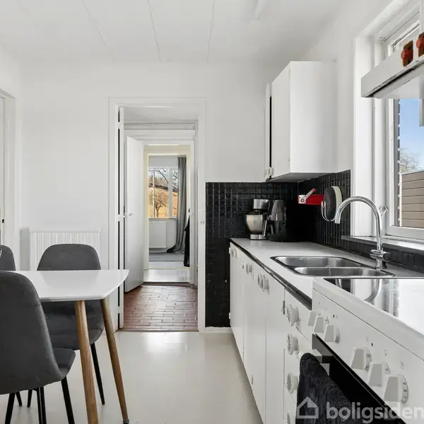 Køkkenbord med grå stole står ved siden af et hvidt køkkenbord mod sorte fliser. Sollys skinner ind gennem vinduet, og en bananklynge ligger på bordet.