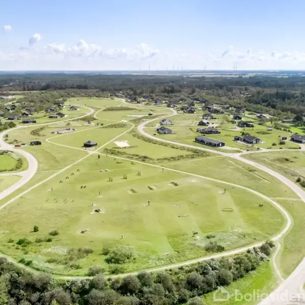 Golfbane set fra oven, flerfarvede huse spredt blandt grønne fairways. Snoede veje forbinder området, omgivet af frodige træer og åbent landskab under en klar himmel.