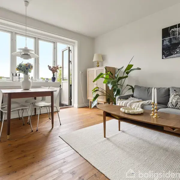 Sofa med puder står ved træbord i stue. Spisebord med stole er ved siden af vinduer med udsigt. Planter og dekorationer skaber en hyggelig atmosfære. Ingen tekst fundet.
