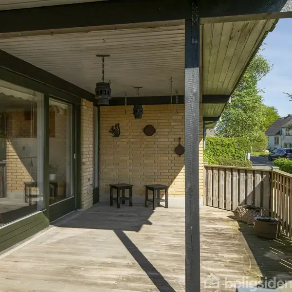 Terrasse med trædæk og rektangulære borde. To lamper hænger fra loftet. Omgivet af et lavt hegn, med udsigt til en grøn have og et nabolag med huse.