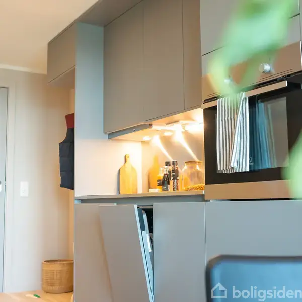 Køkkenskabsvæg med indbygget ovn; håndklæder hænger fra ovnen. Flere flasker og en gul flaske på en oplyst hylde. Moderne design og grå skabslåger.