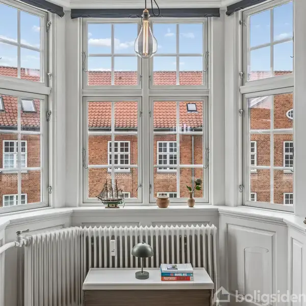 En vindueskarnap med udsigt til røde murstensbygninger. Et lille bord med en bordlampe og bøger foran radiator. Skib og planter i vindueskarmen skaber en hyggelig atmosfære.