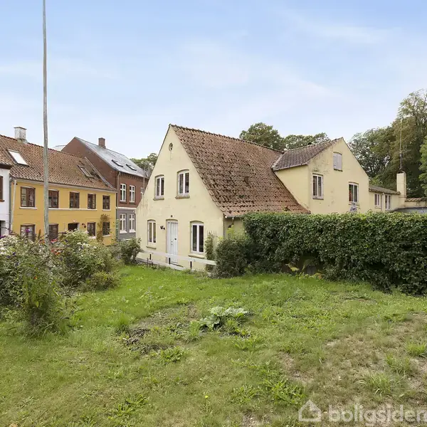 Et gult hus med rødt tegltag står på en græsplæne omkranset af hæk, i en gade med farverige huse. Skoven er i baggrunden under en klar himmel.