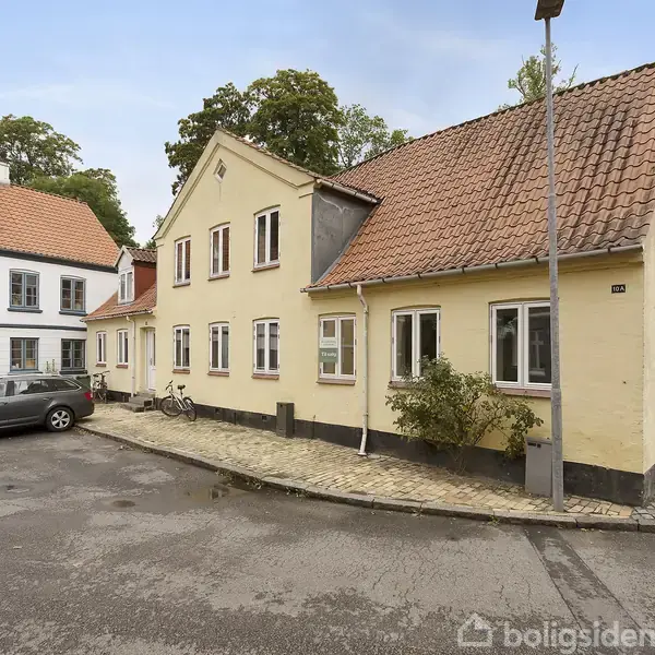 Et gult murstenshus står på en brostensbelagt vej med et rødt tegltag og flere vinduer. En bil er parkeret ved siden af, og træer omgiver området. Husnummeret er 34.
