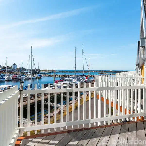 Balkon omgivet af hvidt rækværk strækker sig ud mod marinaen, hvor sejlbåde ligger fortøjet ved kajen under en klar blå himmel.