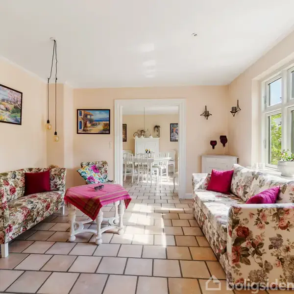 Blomstrede sofaer er placeret omkring et bord med en pink dug; rummet er lyst med store vinduer og vægdekorationer, mens en dør fører til en spisestue i baggrunden.