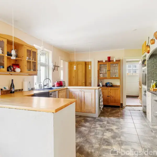 Et rummeligt køkken med lyse træskabe og kogeø, hvor køkkenredskaber og flasker er placeret. Flisegulv strækker sig gennem rummet, der har lyse vægge og et stort vindue.