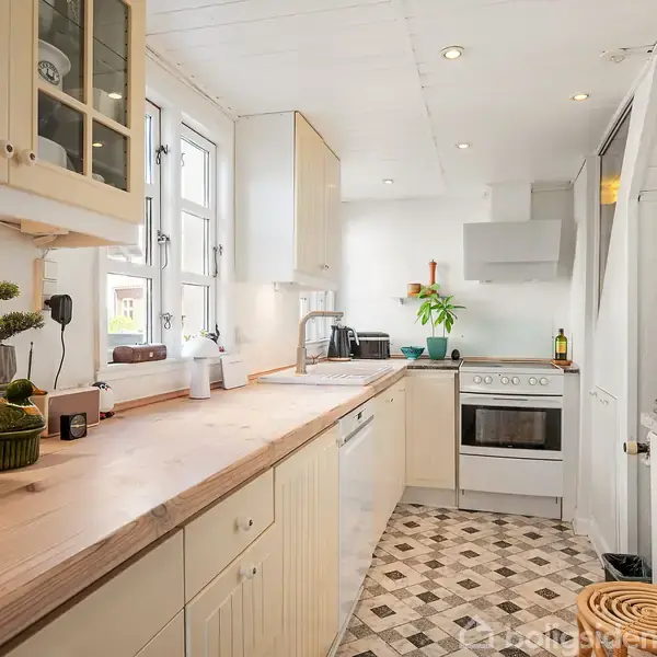 Køkken med træbordplade, hvidt kabinet og moderne apparater, herunder ovn og mikroovn. Der er en plante ved siden af komfuret. Flisegulvet har et mønster, og store vinduer giver lysindfald.