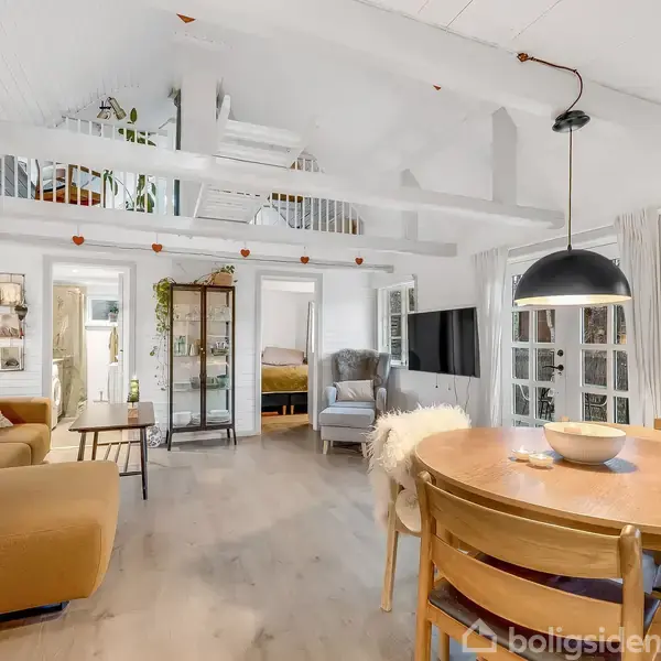 Stue med lyse vægge og træbjælker, indrettet med en gul sofa og rundt spisebord. En trappe fører til en åben hems. Der ses planter og kunst på væggene.