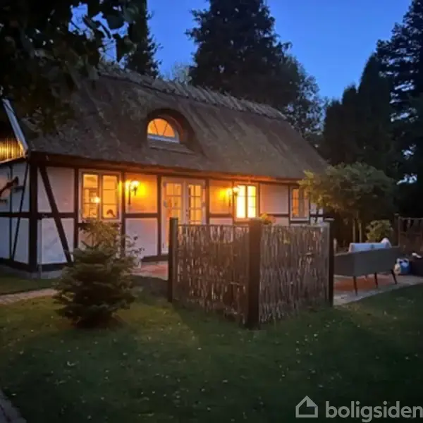 Et bindingsværkshus med stråtag står stille i en grøn have. Stien af sten fører til en lille terrasse med stole. Himlen er blå med skyer.