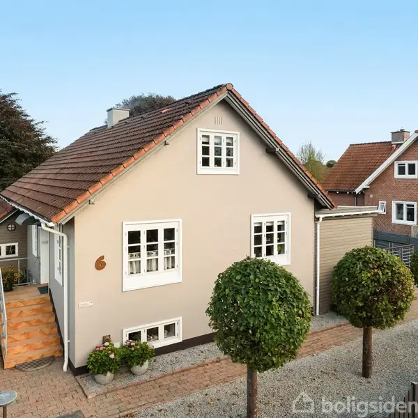 Et ældre, beige hus med rødt tegltag har tre små træer foran sig. Der er en stentrappe ved indgangen. Huset ligger i et villakvarter med nabohuse synlige.
