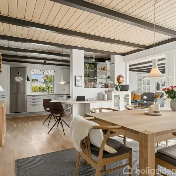 Spisebord med stole står i centrum af en lys stue med trægulv og åbent køkken. Store vinduer og hylder med dekorative genstande skaber en hyggelig atmosfære.