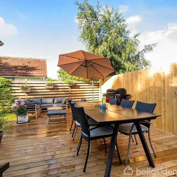 En træterrasse har et spisebord med stole, en parasol og en grill. Konteksten er en hyggelig have med blomster og et træhegn ved siden af et hus.