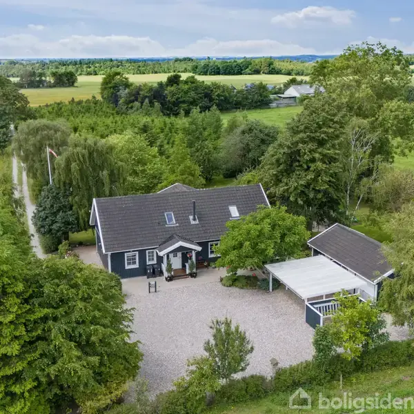 Et sort hus med hvide vinduesrammer står i midten, omgivet af grønne træer og buske. En grusvej fører til huset, der ligger i landlige omgivelser.