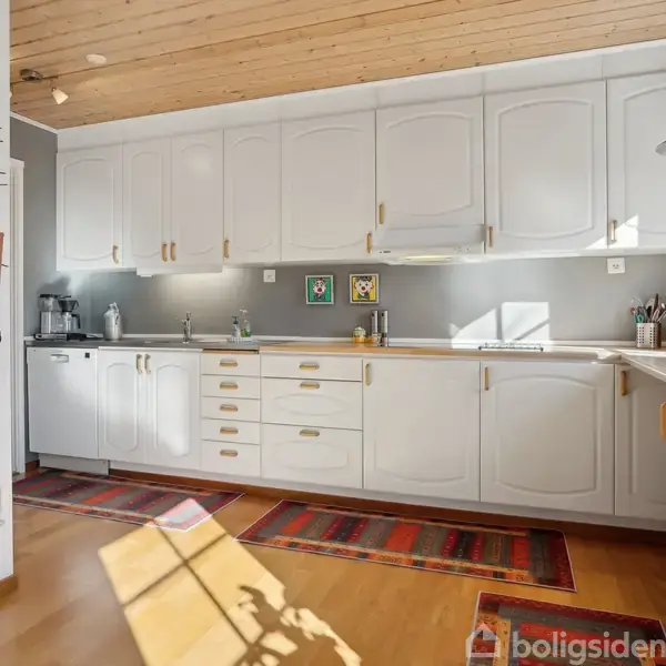 Et køkken med hvide skabe og indbyggede apparater, herunder en ovn. Sollys skinner på trægulvet. Der hænger en opslagstavle med viskestykker på væggen. Tre farverige tæpper ligger på gulvet.