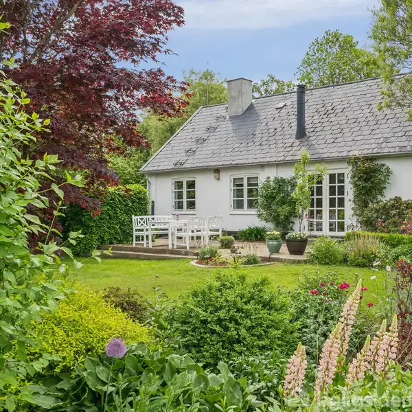 Hvidt hus med gråt tag står i en frodig have. Terrasse med hvide havemøbler foran huset. Træer og buske omgiver området, der emmer af rolig, landlig idyl.