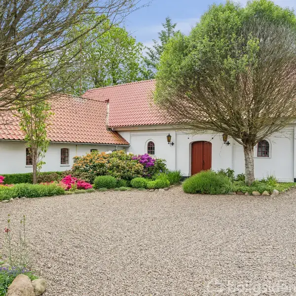 Et hvidt hus med rødt tegltag står blandt farverige blomster og grønne buske, omkranset af en grusbelagt gårdsplads og træer i en fredelig haveomgivelse.