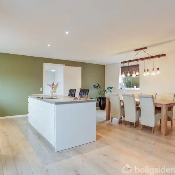 Et soveværelse med en dobbeltseng dækket af en dyne, placeret ved siden af et sengebord med lampe. En kommode står ved døren, der fører til et badeværelse.