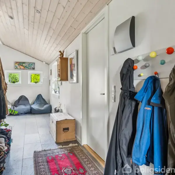 Gang med tøj på farverige knager, rød løber, to sorte sækkestole under små vinduer og en hylde med dekorative genstande. Lyse trævægge og loft giver en lys atmosfære.