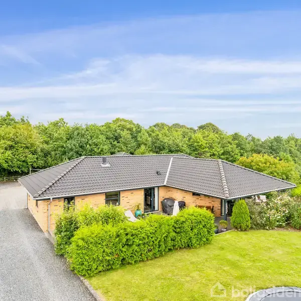 Et gult murstenshus med sort tag står omgivet af grønne hække og en grusvej, med en baggrund af frodig skov og åbne marker under en blå himmel.
