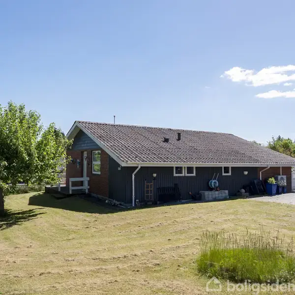 En etplanshus med mørkegrå vægge står stille på en velplejet græsplæne. Huset ses fra siden, med en terrasse og havemøbler i nærheden. Himlen er klar med få skyer.