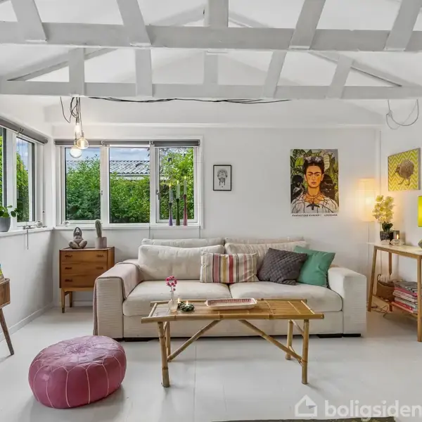 Stue med hvid sofa fyldt med puder står mod en væg med kunst og en lampe. Et sofabord foran, et skuffemøbel til venstre, grønne planter udenfor.